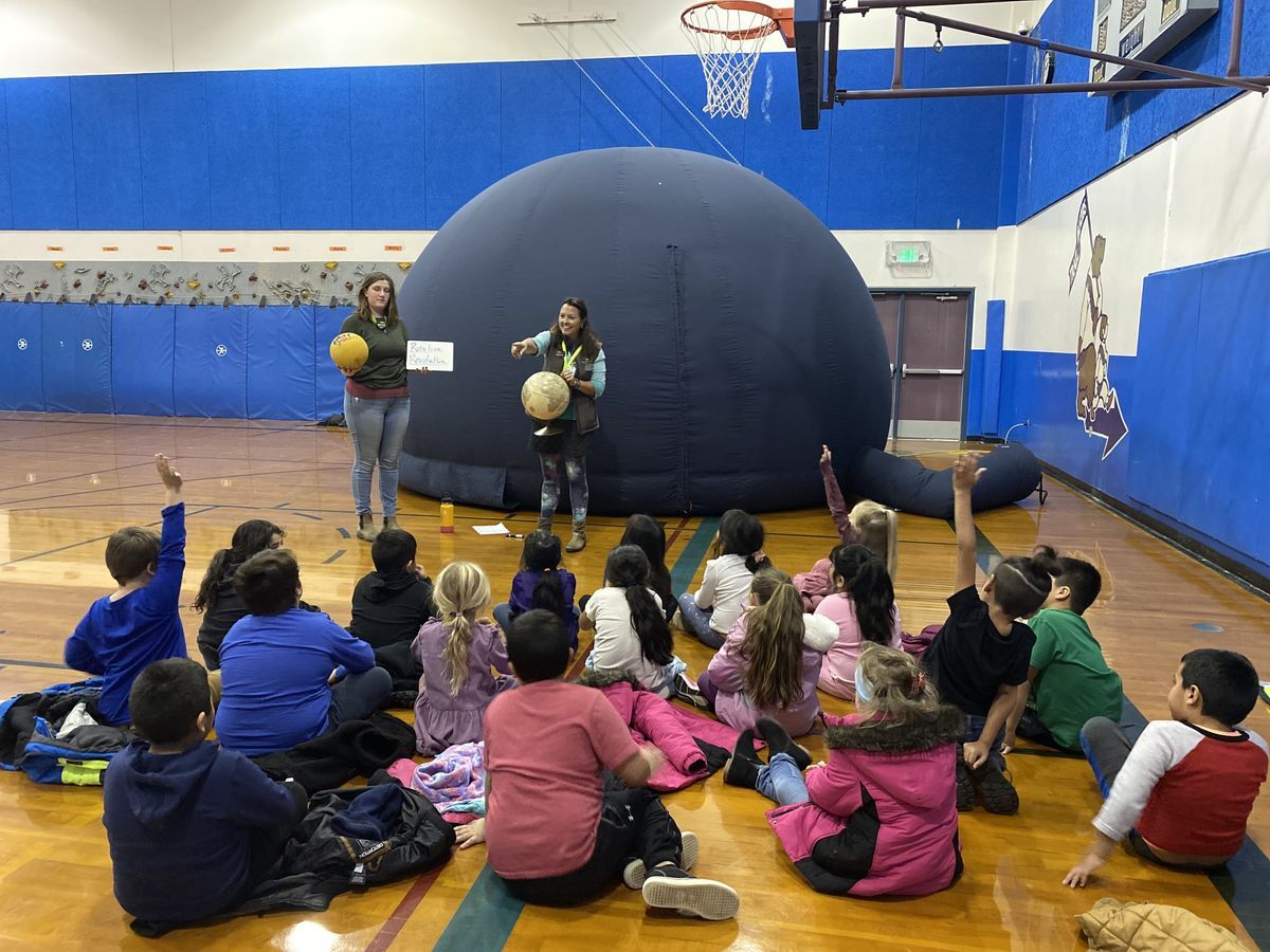 Two staff holding a globe standing in front of the large black domed planetarium speaking to a group of students.