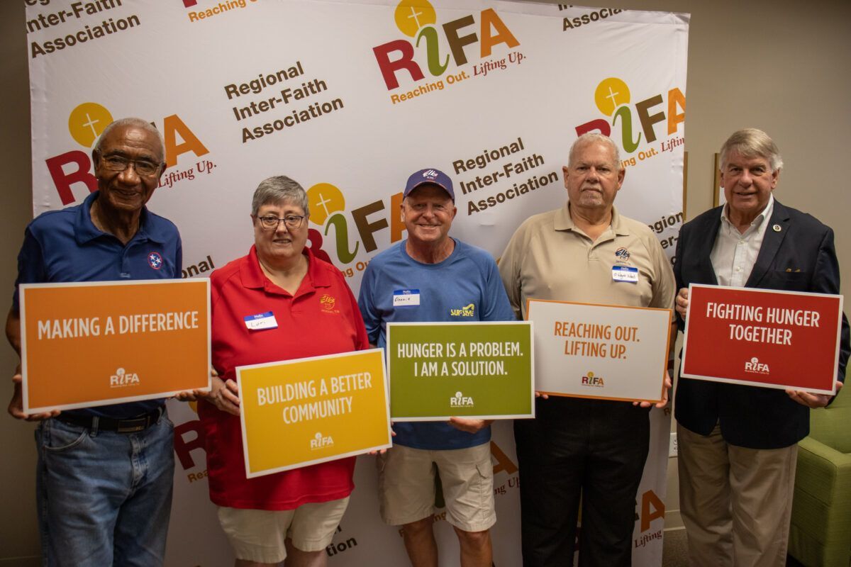 Five people holding signs with messages about community and hunger relief in front of a Regional Inter-Faith Association backdrop.