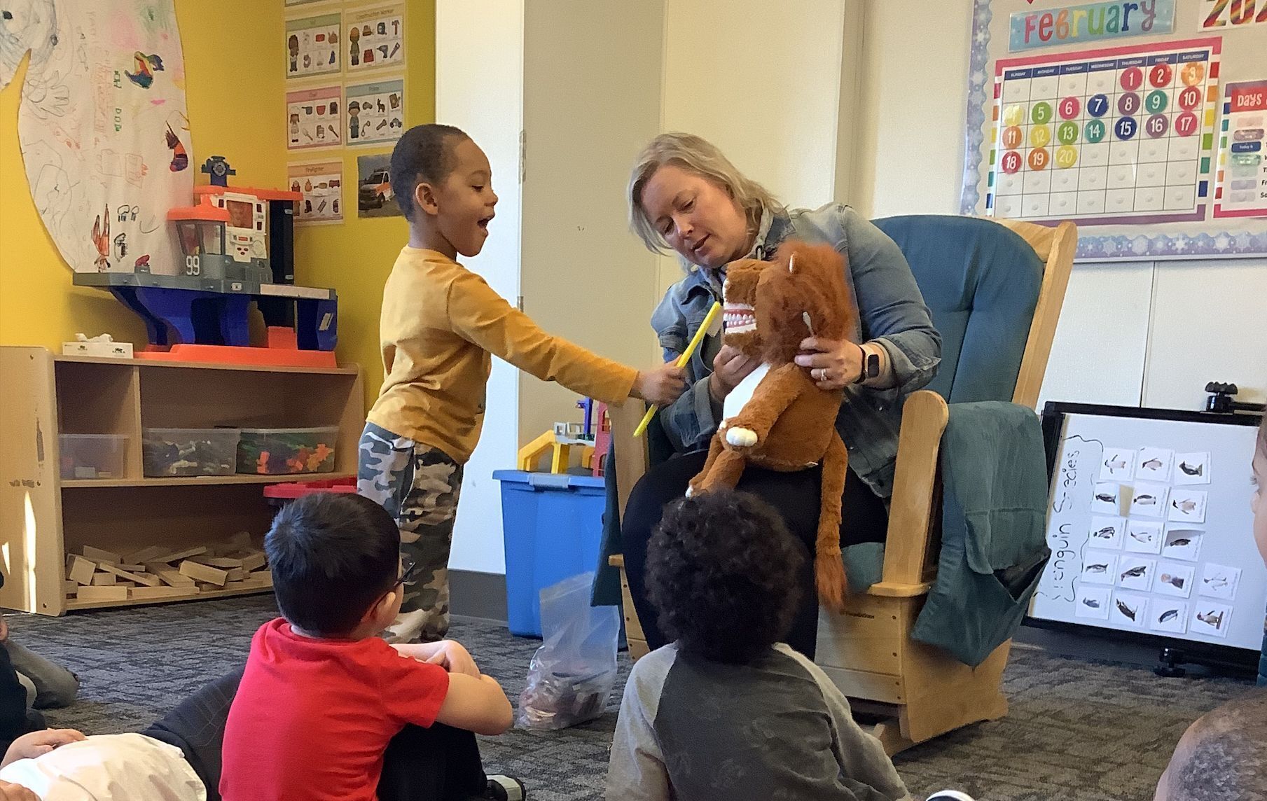 A lady is holding an animal puppet that is showing big fake teeth, while a preschool student "brushes its teeth" with a toothbrush. About a half dozen other preschoolers sit and watch.