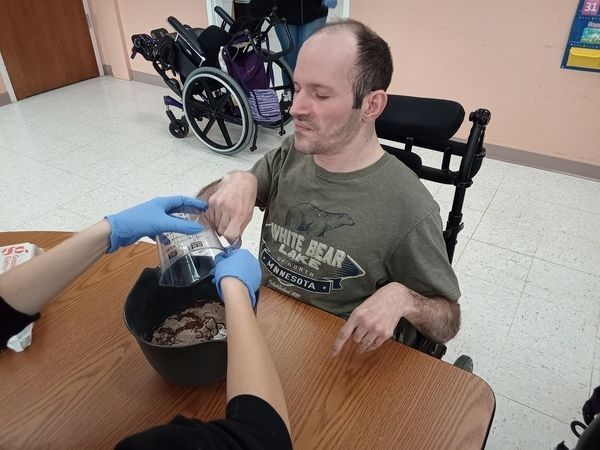 A man in a wheelchair dumping a liquid into a mixing bowl during a culinary class. 