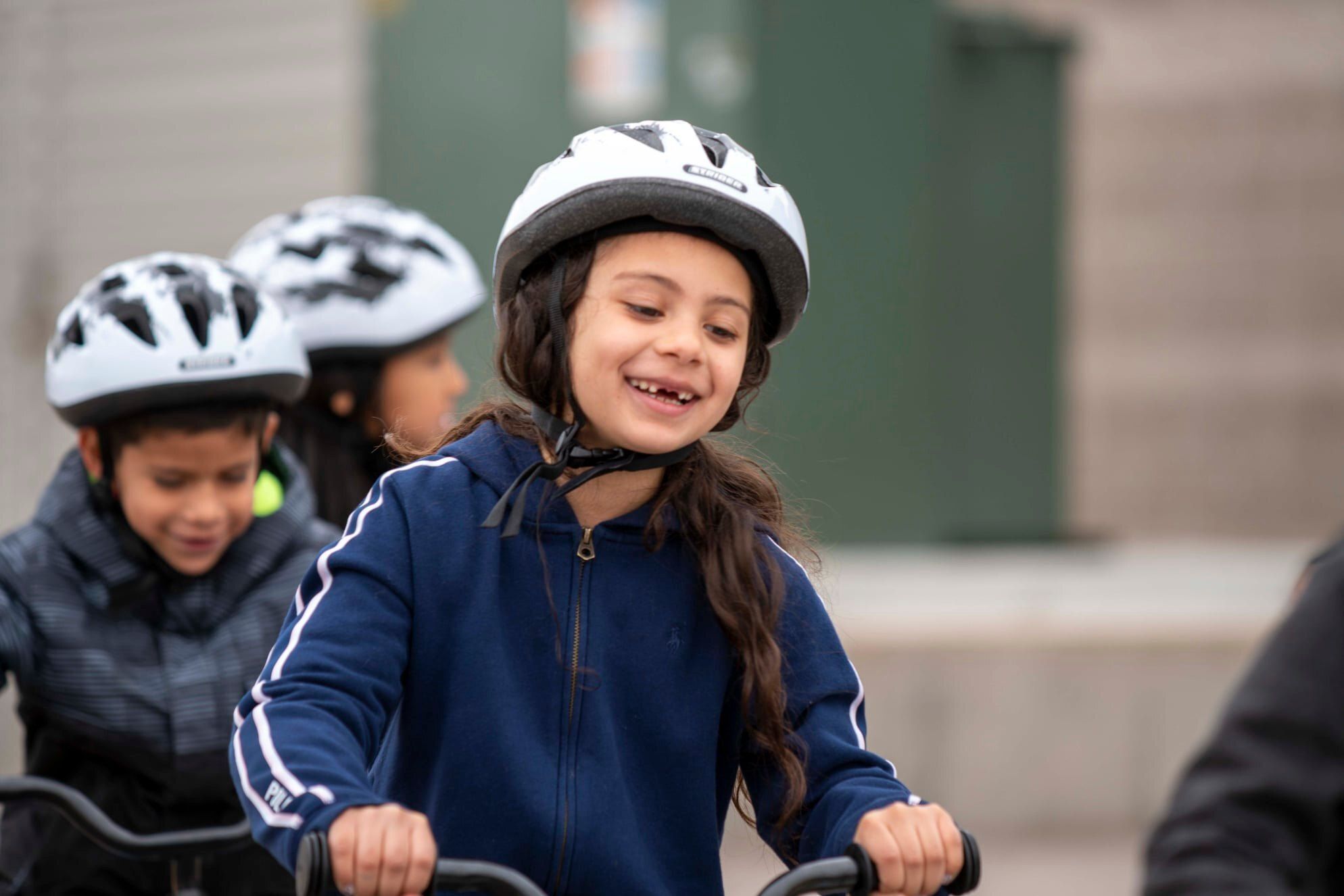 A young girl smiles as she rides her bike, wearing a helmet and navy jacket, with other children biking beside her.  The moment captures confidence, learning, and the joy of new experiences.