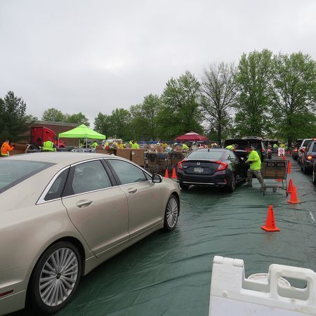 This is an image of a community hazardous household waste disposal event. There are cars lined up to drop off items at tents and many volunteers in safety vests are walking around.