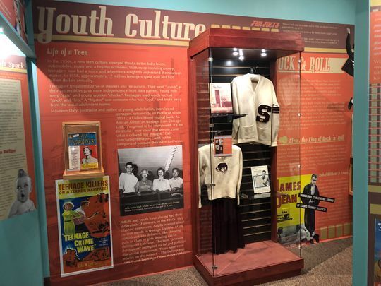 Exhibit labels and case at the Smoky Hill Museum about 1950 teen culture. Items on display are a Salina Central High School letterman sweater and uniform.