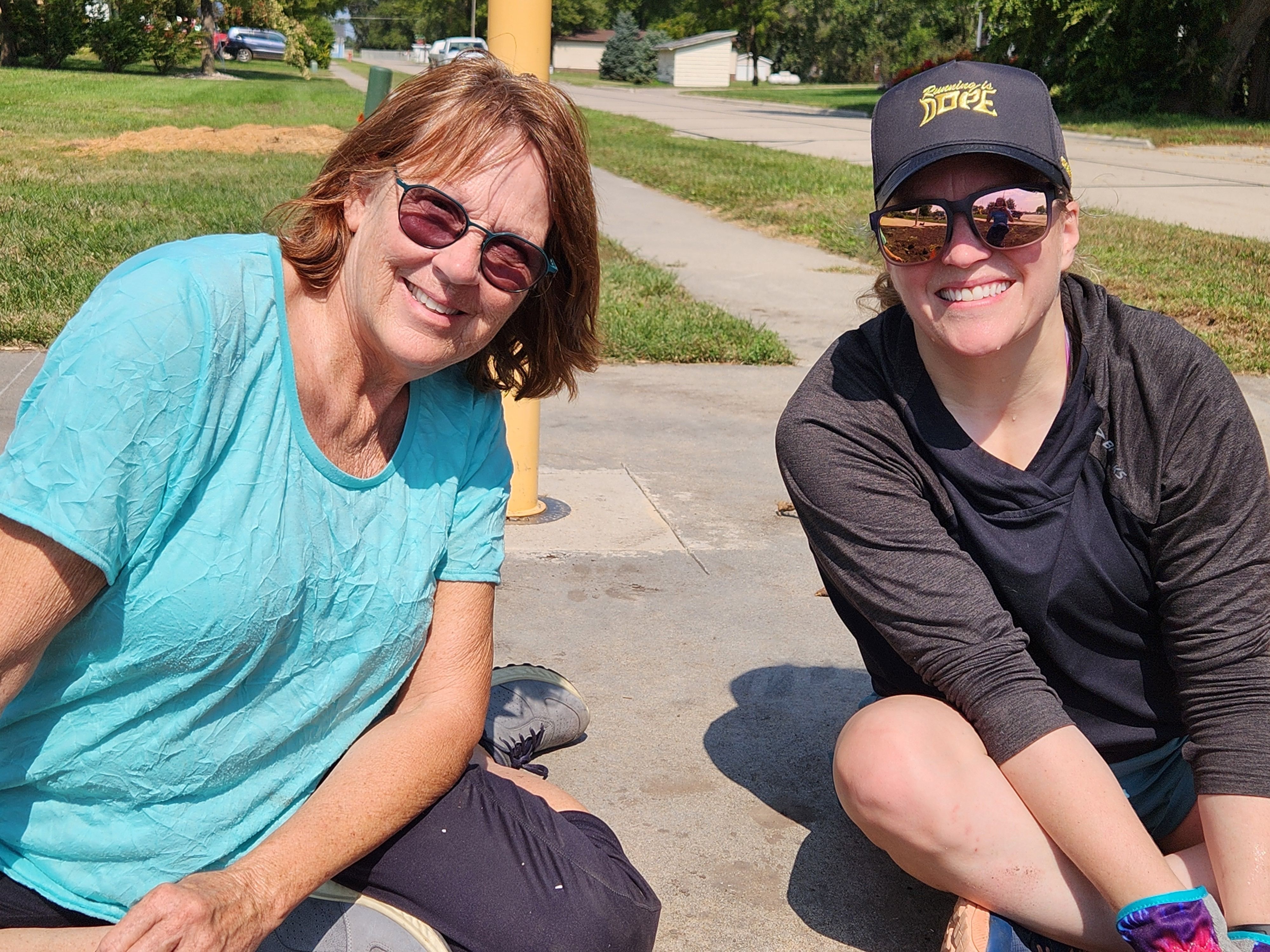 Two ladies sitting on a sidewalk planting perennials in a dirt bed. 