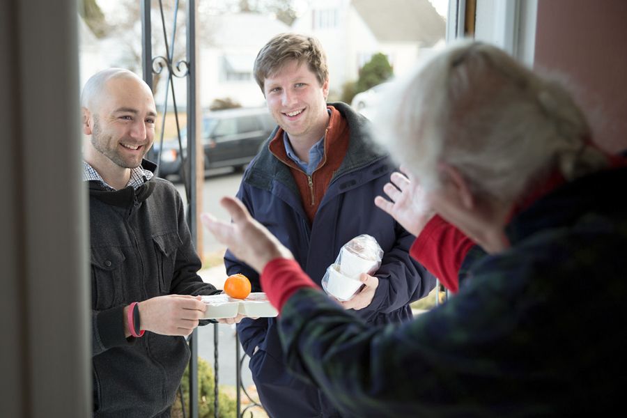 Two young men delivering food for Meals on Wheels to older woman who is welcoming them at her front door