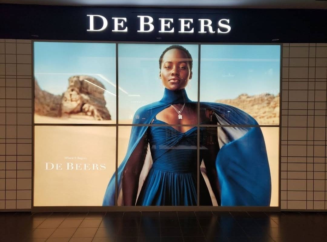 High-end backlit retail display featuring the De Beers logo and a vibrant, illuminated promotional image of a woman in a royal blue dress wearing diamond jewelry, installed in a tiled mall wall.