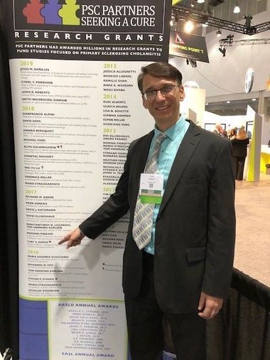 A man is pointing to his name on a PSC Partners standing sign. He is wearing a dark suit and smiling at the camera.