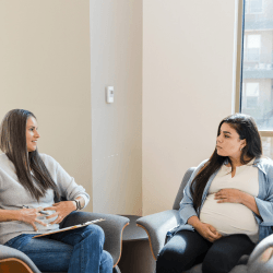 A pregnant mother and her mentor sit in a bright open room on couches, talking animatedly.