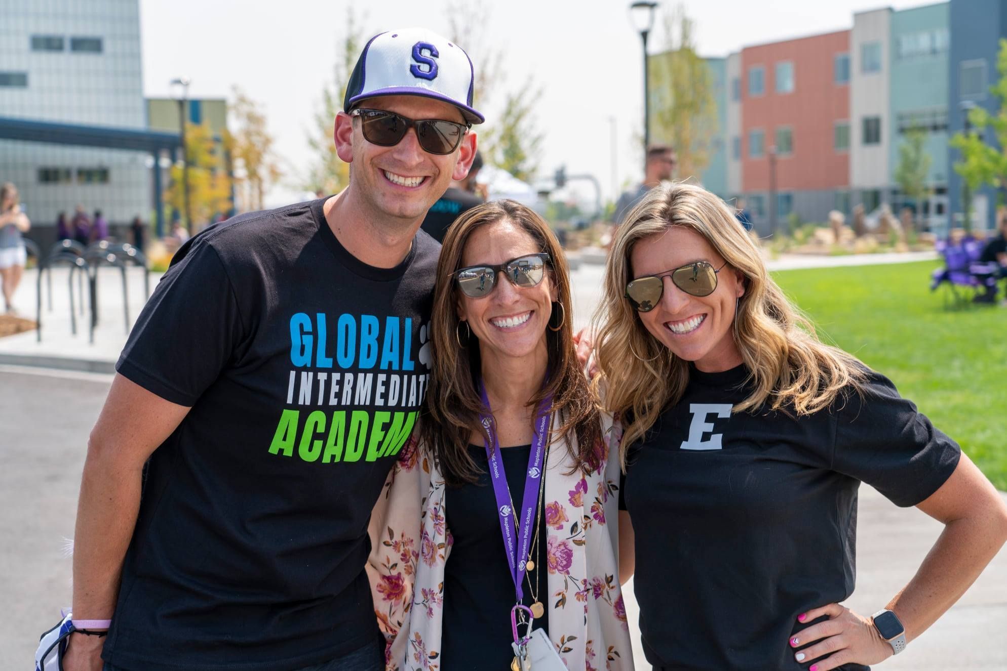 Three smiling adults in school t-shirts pose outdoors on a sunny day, representing the collaborative spirit of Mapleton Education Foundation Chapters.