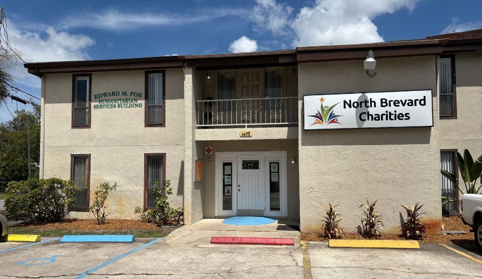 Photo of the exterior of North Brevard Charities' client services building. It's tan with dark brown trim, parking spots out front, and a branded "North Brevard Charities" sign to the right on the building.