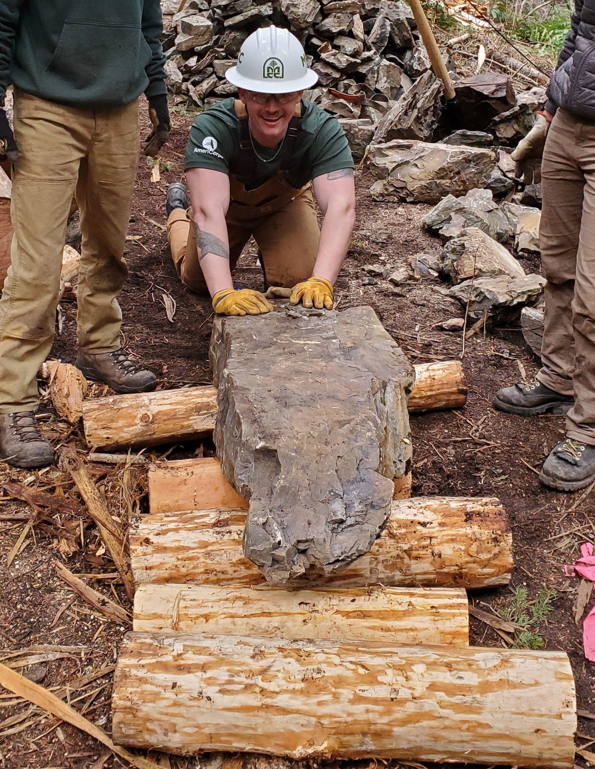 A crew leader wearing a helmet, gloves, and glasses rolls a large flat rock over several logs to transport it down trail.