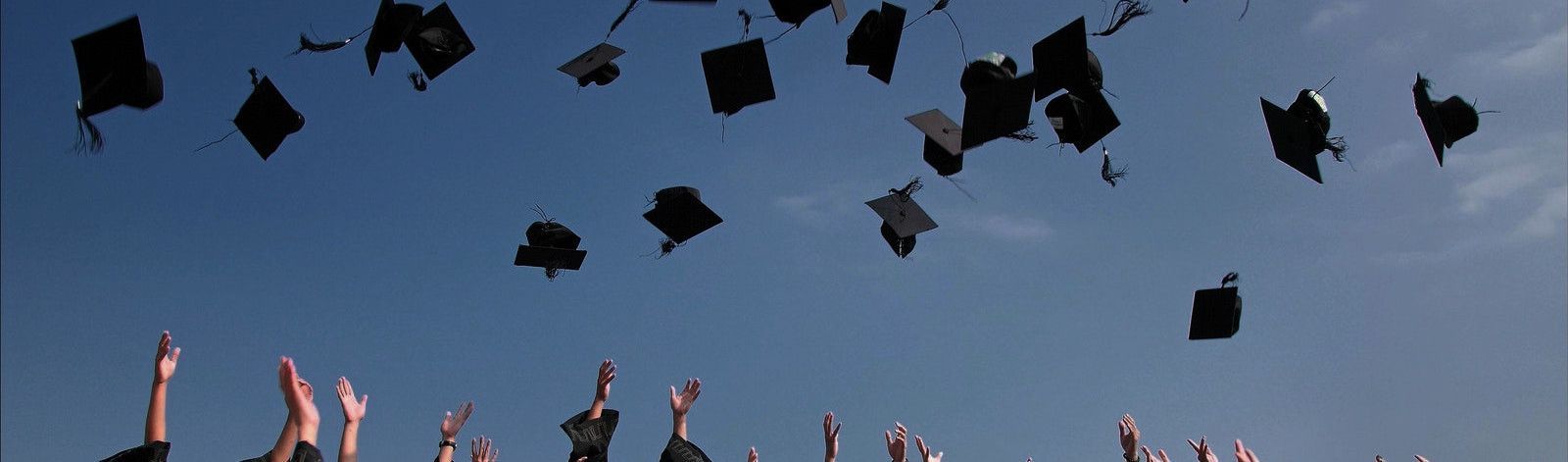 Photo of graduate students in black gowns throwing their matching mortarboards in the air. The mortarboards are suspended in the air at the top against a blue sky. Student hands point upward from the bottom of the photo as they throw their hats in the air