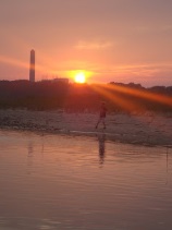 This is a picture of a sunset on the beach. A person is walking on the beach, and a tall building is in the distance.