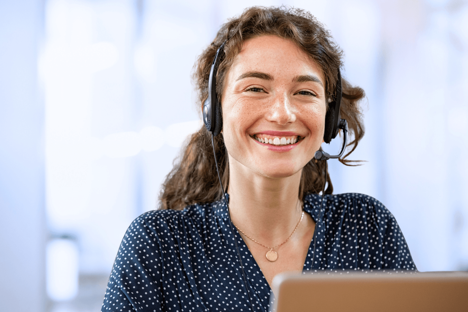 Brunette HelpLine advocate with freckles and curly hair smiles while wearing a hands-free phone headset. She is seated at her computer in a bright office.