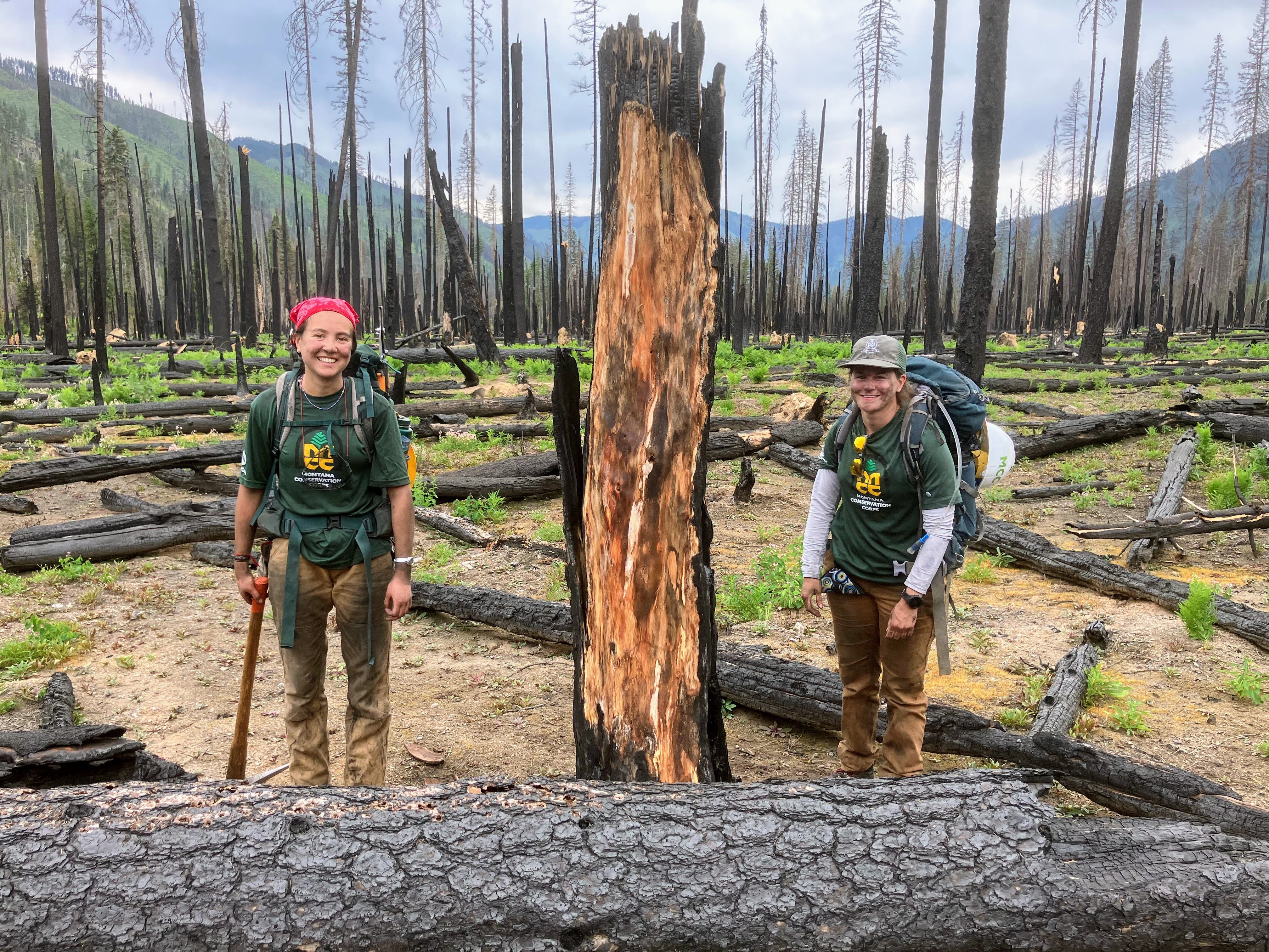 Two crew members stand next to a charred log across the trail, smiling.