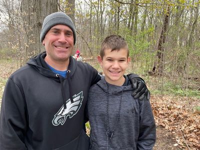 A Father wearing an Eagles sweatshirt hugs his son while facing the camera with a pile of leaves in the background