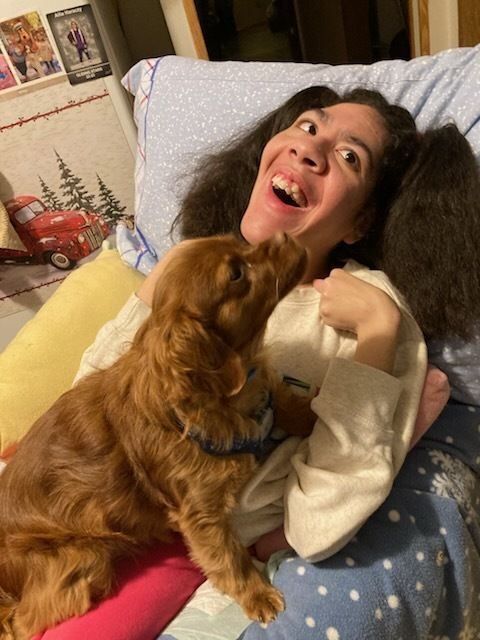 A young girl is laying on a bed and laughing while a dog rests on her chest.