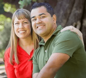 A smiling couple is outdoors with the woman wearing a red shirt and the man wearing a green polo shirt. The woman has her arm around the man. The background features a large tree and soft natural light creating a warm and relaxed setting.