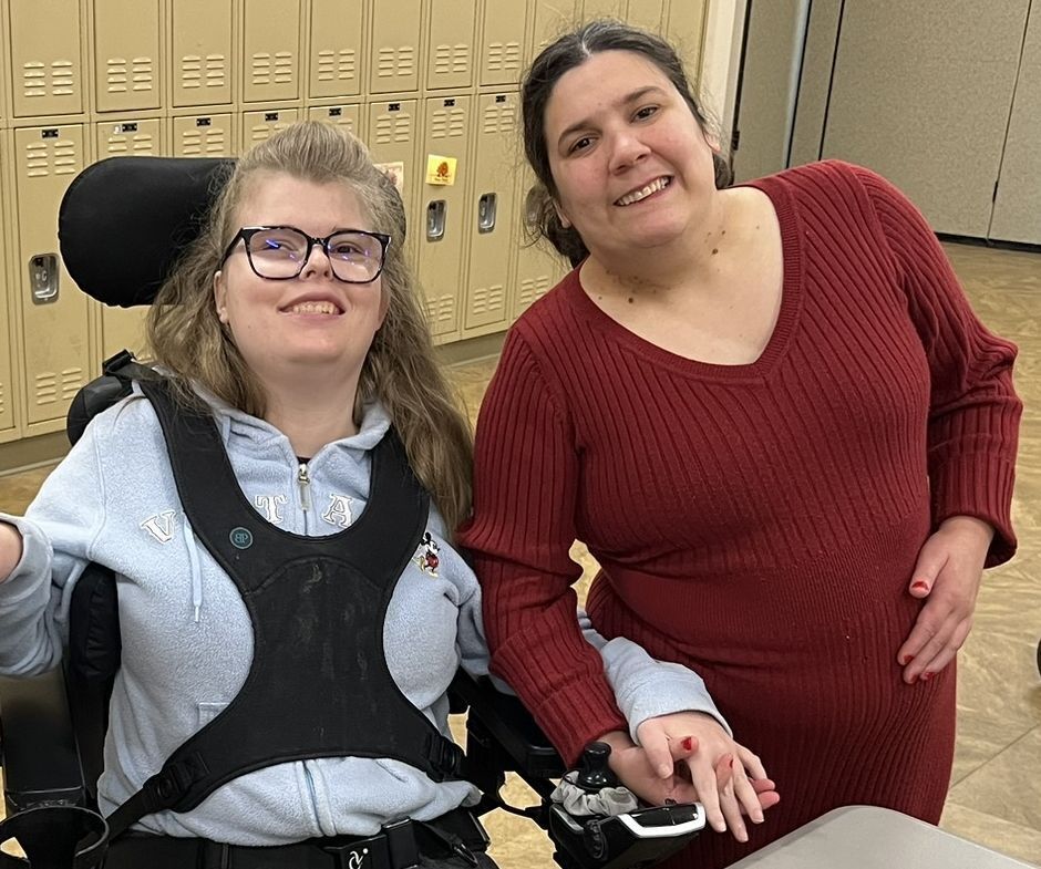 A woman stands smiling next to a woman sitting in a wheelchair, also smiling.  They are holding hands.