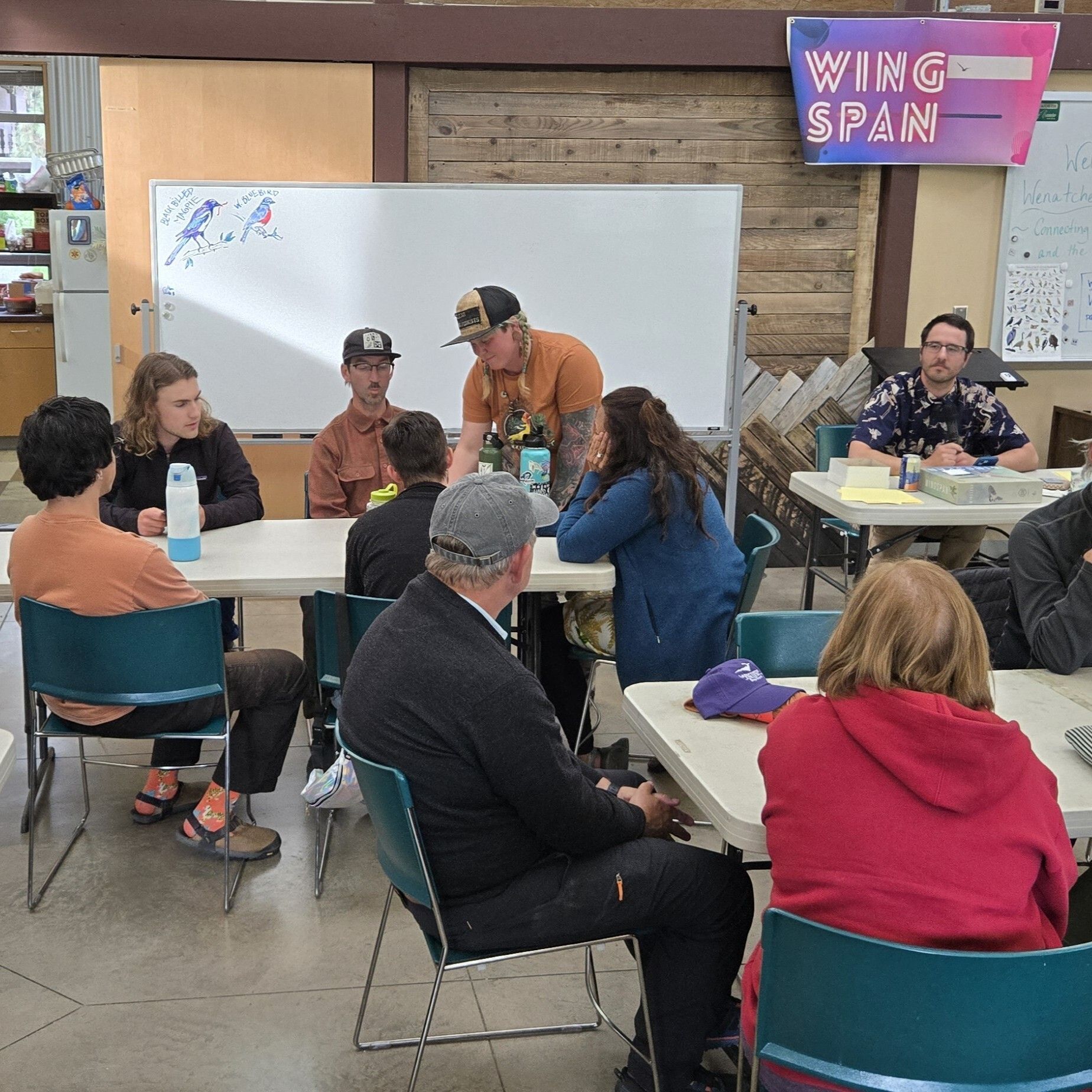 Several people sit at tables, looking at an announcer under a Wingspan game banner