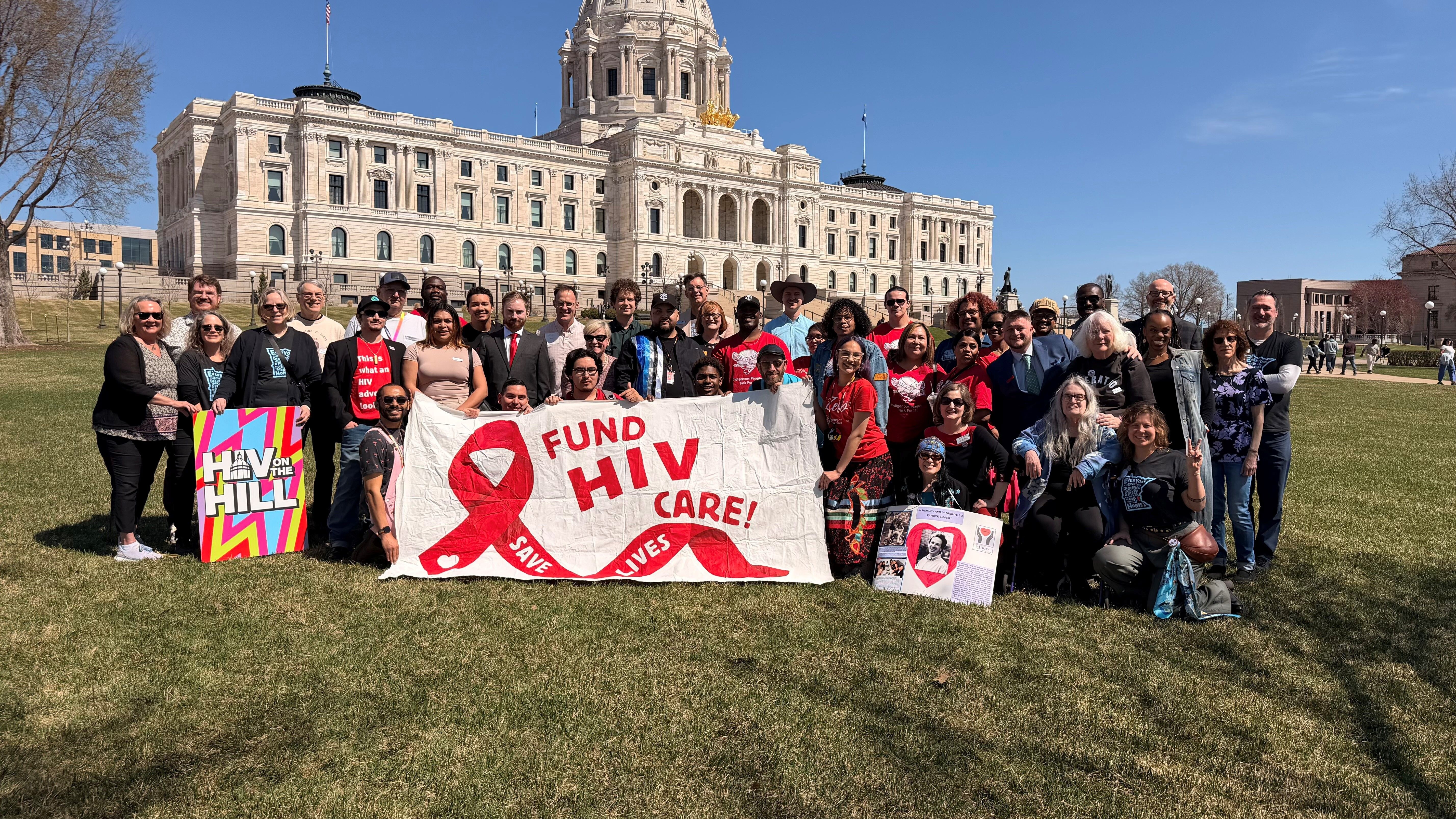 44 people stand on the Capitol Lawn holding a sign that says Fund HIV Care - Save Lives