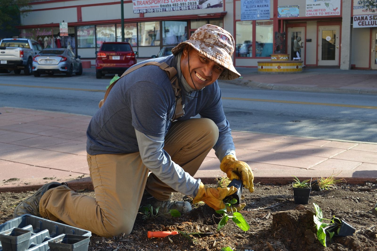 A Latino man wearing a tan hat knees to plant flowers in a raised bed.,