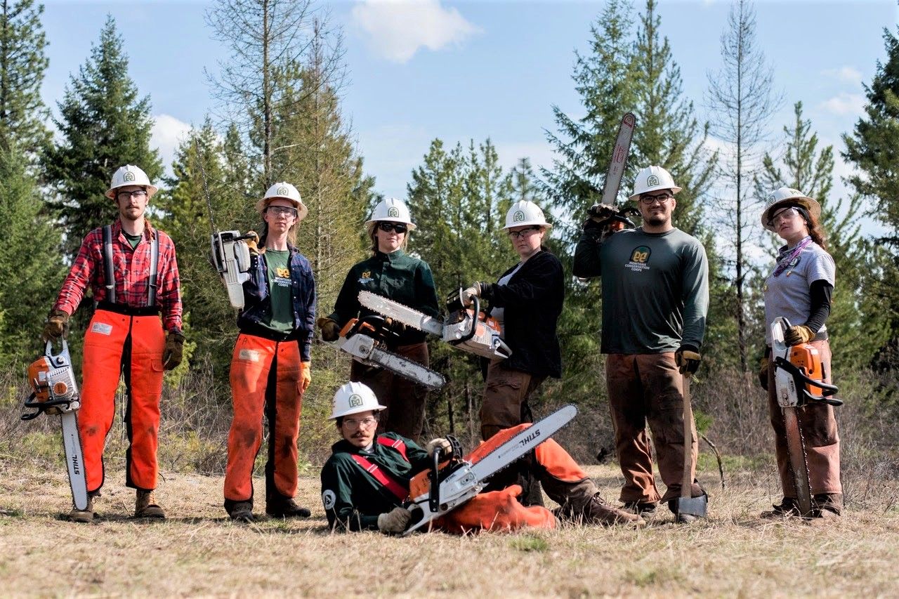 [Image Description: Seven MCC members wielding chainsaws stand together. Three members are wearing bright orange safety chaps but all members are dressed in their MCC uniforms!]