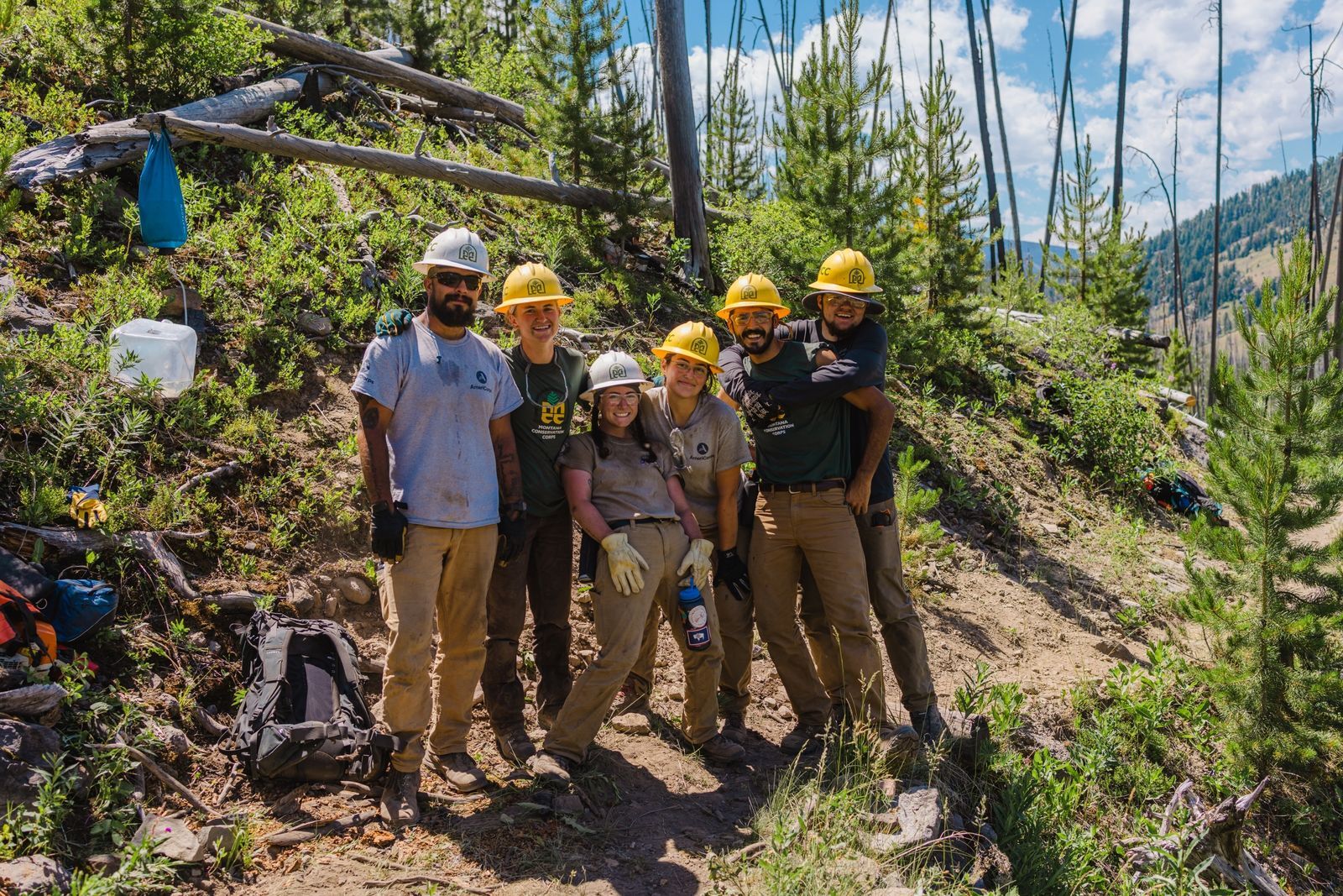 [Image Description: Four MCC members and two leaders pose together on a trail they are working on, smiling and wearing their MCC uniforms and hard hats.]