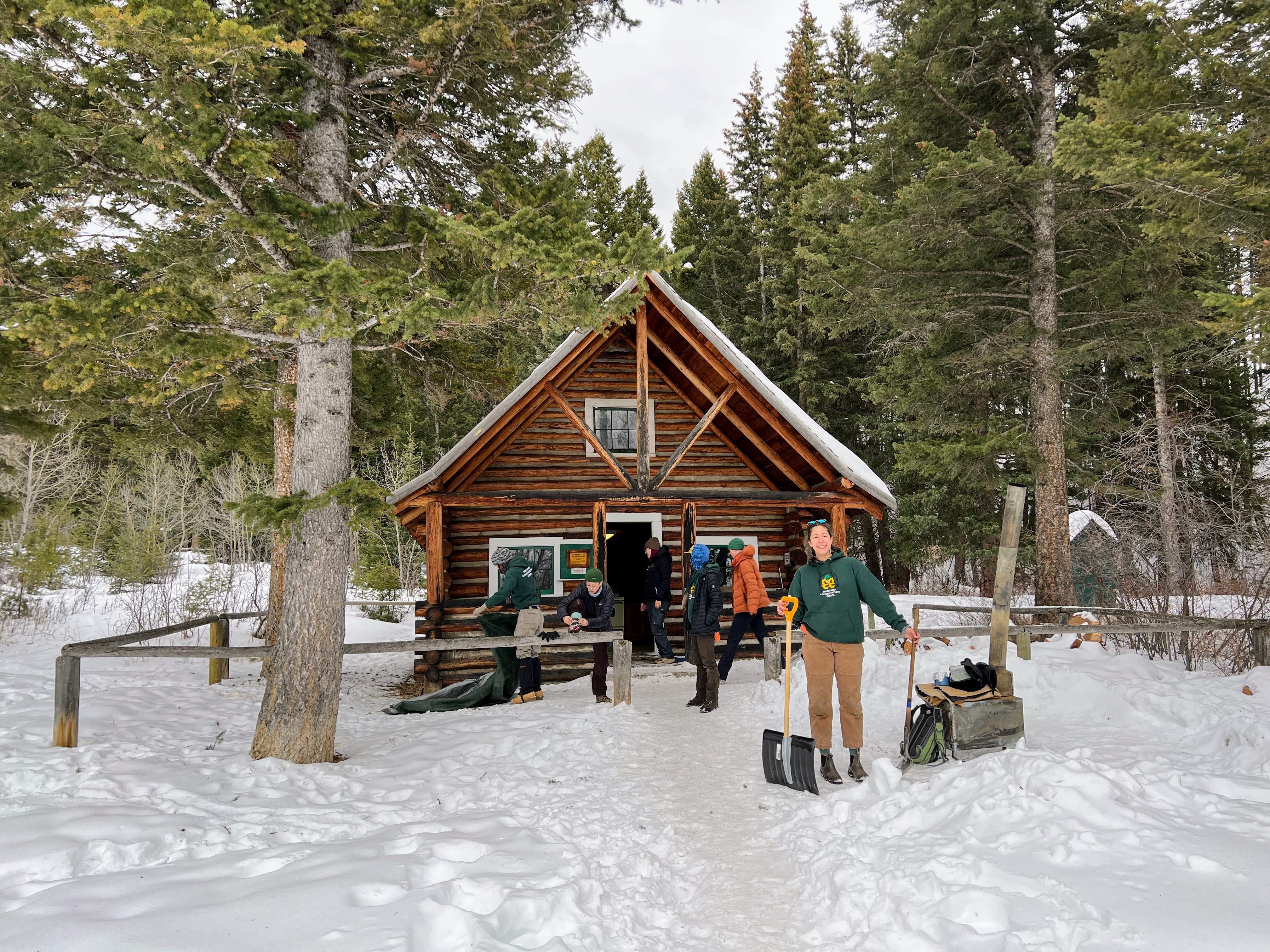 A crew leader holding a snow shovel stands in front of an A-frame cabin. The cabin is surrounded by trees and covered in snow, while other crew leaders work in the background.