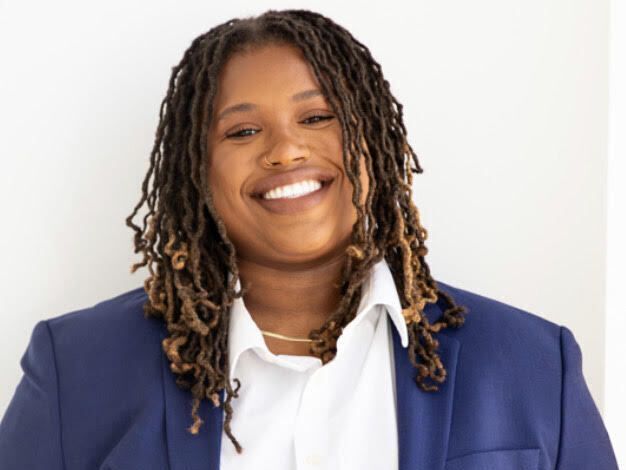 Young Black woman with braided hair parted in the middle and nose ring smiles at camera. She is wearing a blue blazer and white collared shirt.