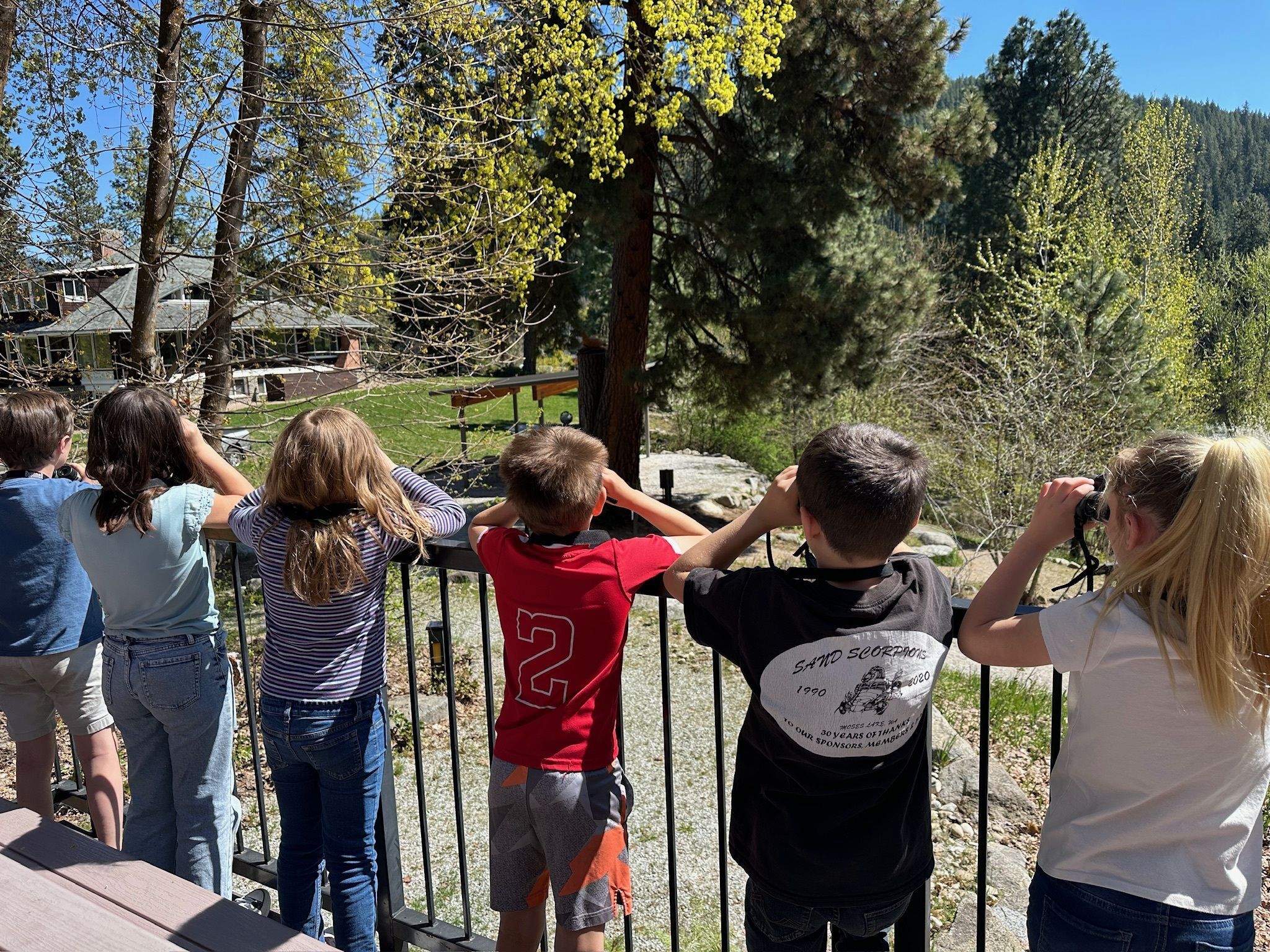 A group of 5 third grade students using their binoculars to spot birds from the Red Barn porch at WRI.