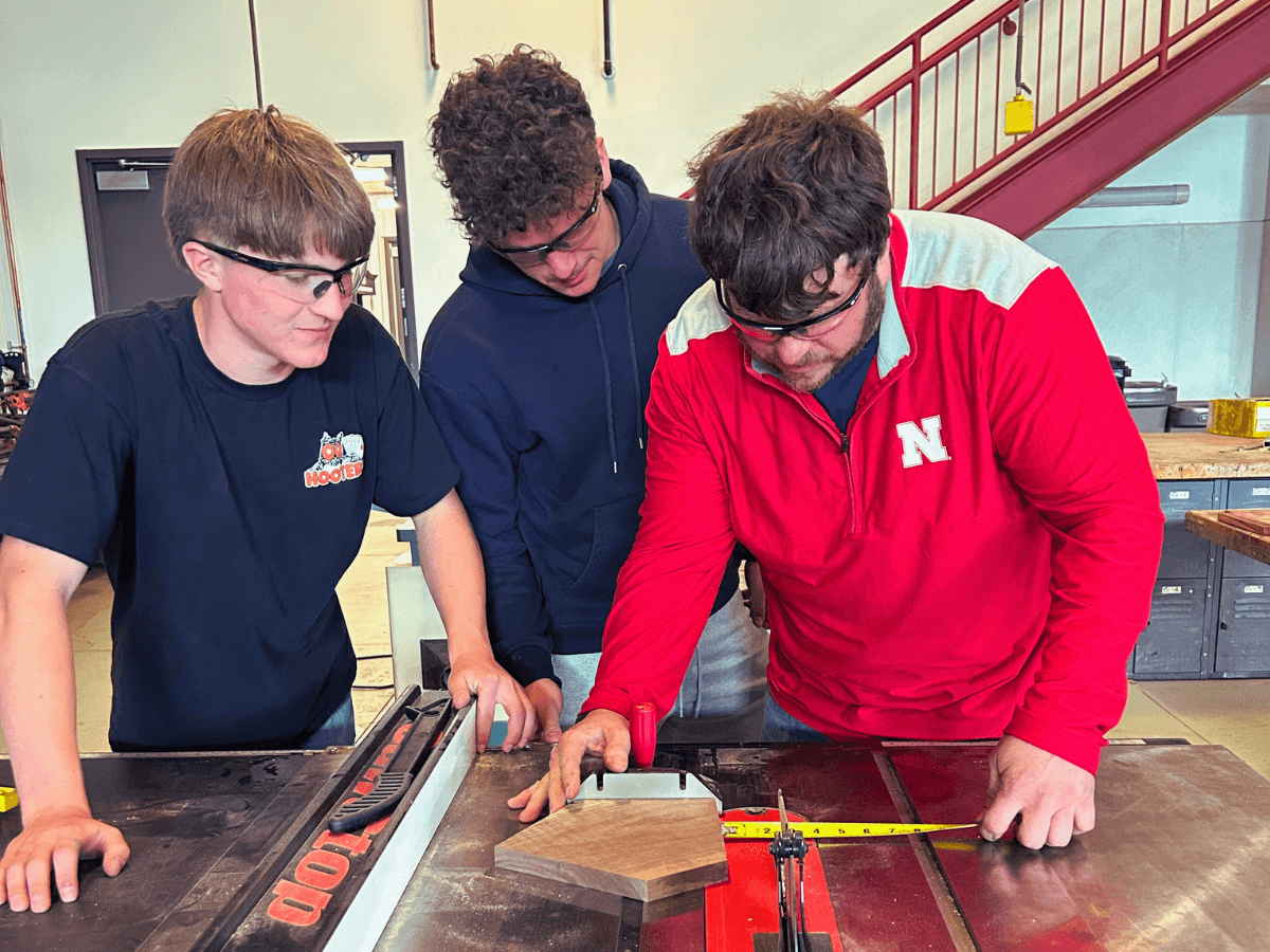 Teacher is helping two students in shop class with the table saw. 