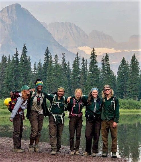 [Image Description: Six crew members arm in arm, all wearing back packing back packs, in front of trees and a beautiful dramatic mountain landscape.]