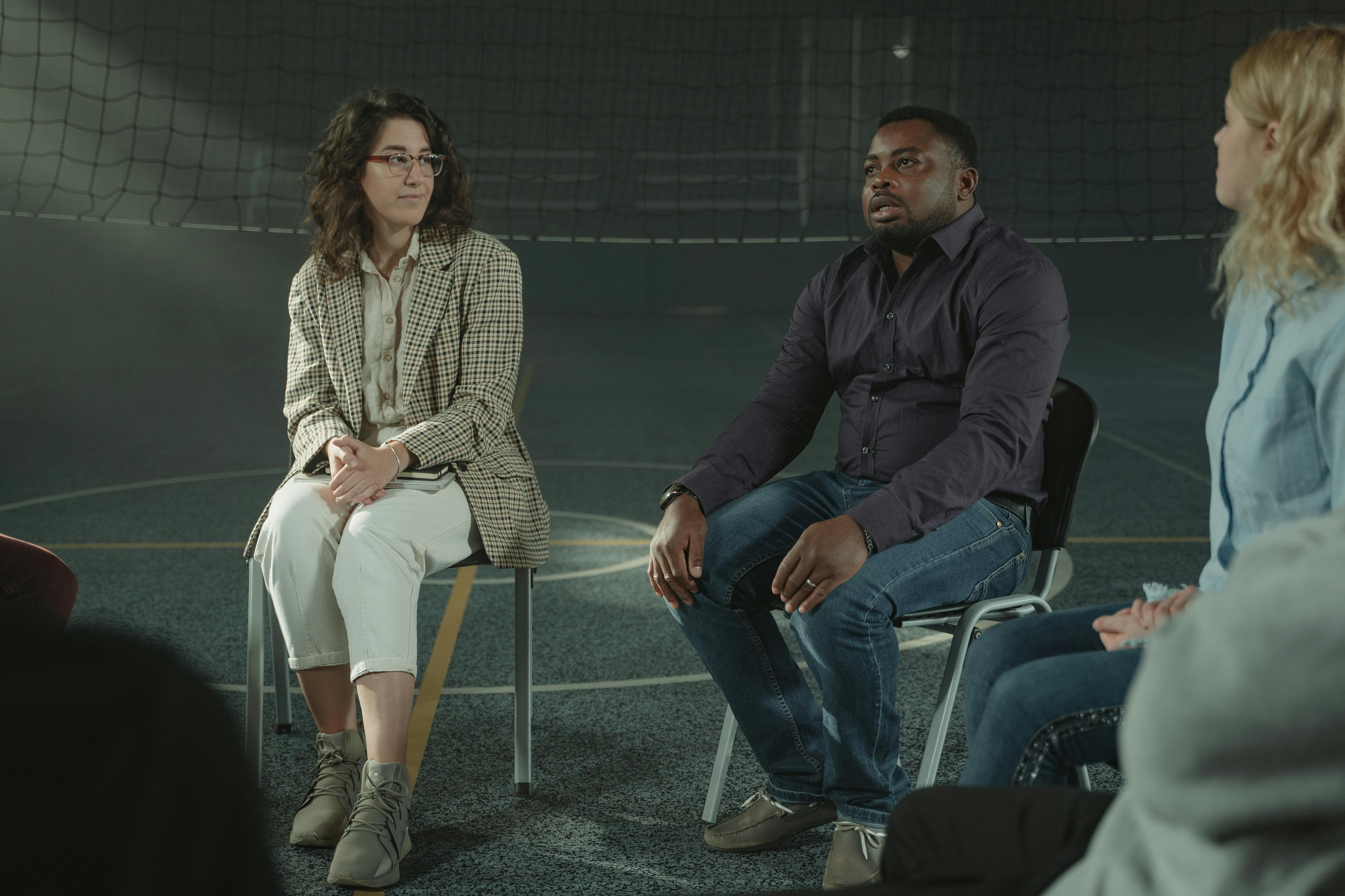 Three people are sitting on folding chairs in a gymnasium listening to an individual in the middle as they speak.