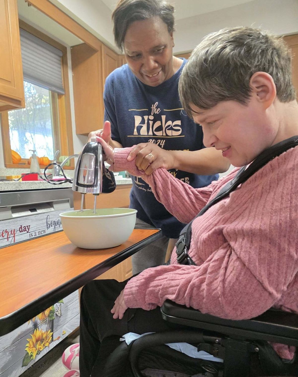 A young woman in a wheelchair faces a countertop while a woman stands to her right helping her hold and operate an electric mixer.