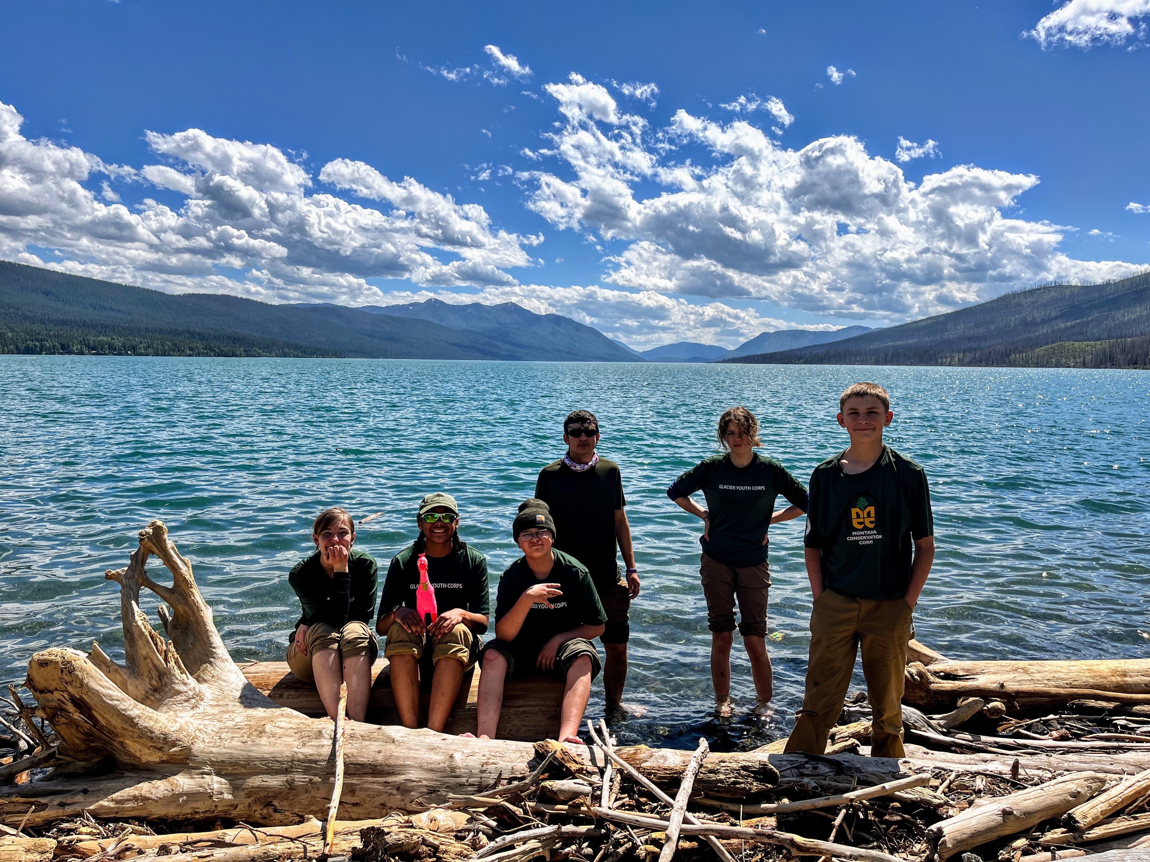A youth crew sits, smiling on logs that border the edge of a large lake. One crew member is holding a pink rubber chicken.
