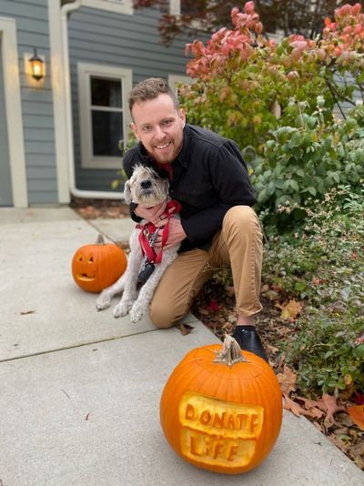 A smiling man is kneeling with his fluffy grey dog. A carved pumpkin is in front of him with the words Donate Life carved on it.