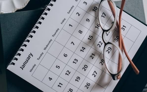 Aerial photo of a monthly calendar, featuring January, at an angle atop a desk. The calendar has a white background with black lettering. A pair of brown reading glasses sit on the right corner of the calendar.