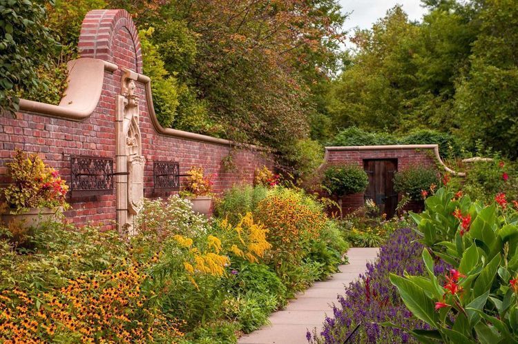 A lush garden pathway lined with colorful late-summer flowers curves alongside a decorative red brick wall with stone accents and wrought iron details. Purple, yellow, orange, and red blooms fill the borders, with dense green trees and shrubs surrounding 