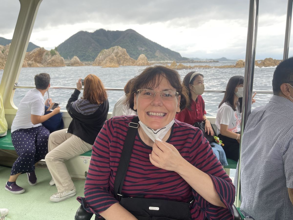 A picture of Ann Marie who’s sitting and wearing a maroon strip sweater and glasses. She has a black hand bag on her shoulder and she pulling her mask down while smiling for the picture. She is on a boat tour in Tottori, Japan.