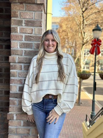 White woman with long blond hair, white shirt and jeans stands smiling by a brick wall. The soft background shows a large tree with golden leaves, and a multi-floored building in the near distance. Flower pots dangle from trees. The sidewalk is brick.