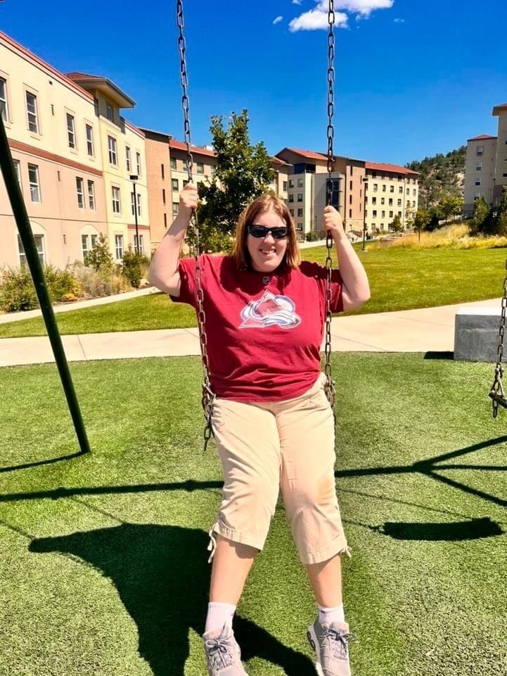 Kaitlin is sitting on a swing on a sunny day, wearing a colorado avs t shirt and smiling. 