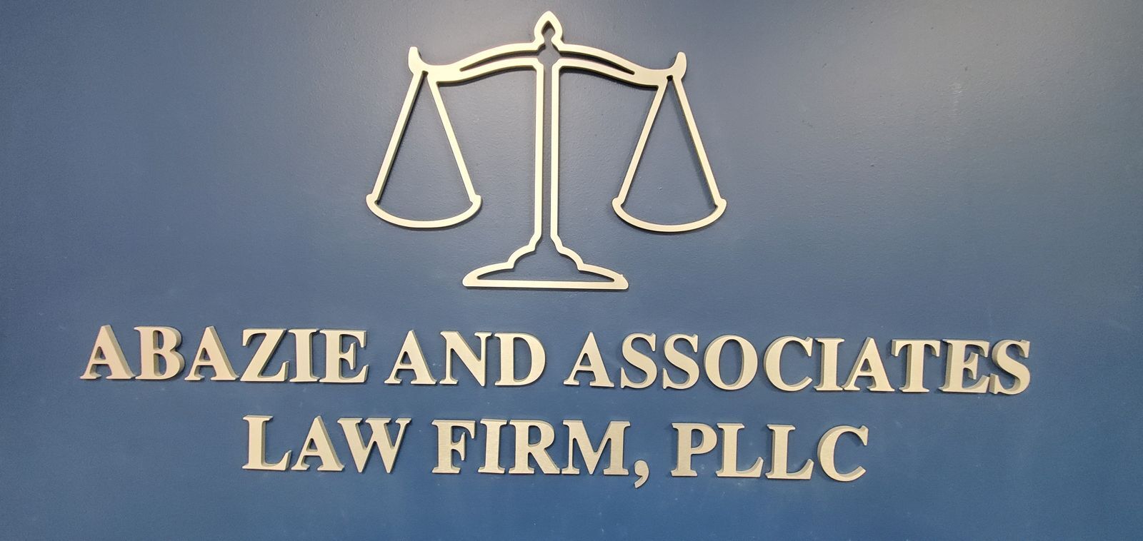 Dimensional lobby signage for “Abazie and Associates Law Firm, PLLC” featuring silver acrylic letters and a scale of justice graphic, mounted on a solid blue background.