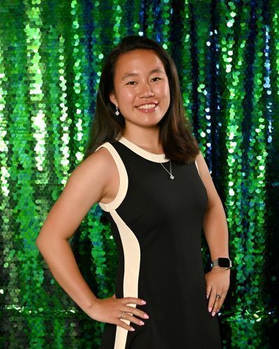 Asian woman with black and white dress stands smiling with hands on hips in front of a sparkling green background.