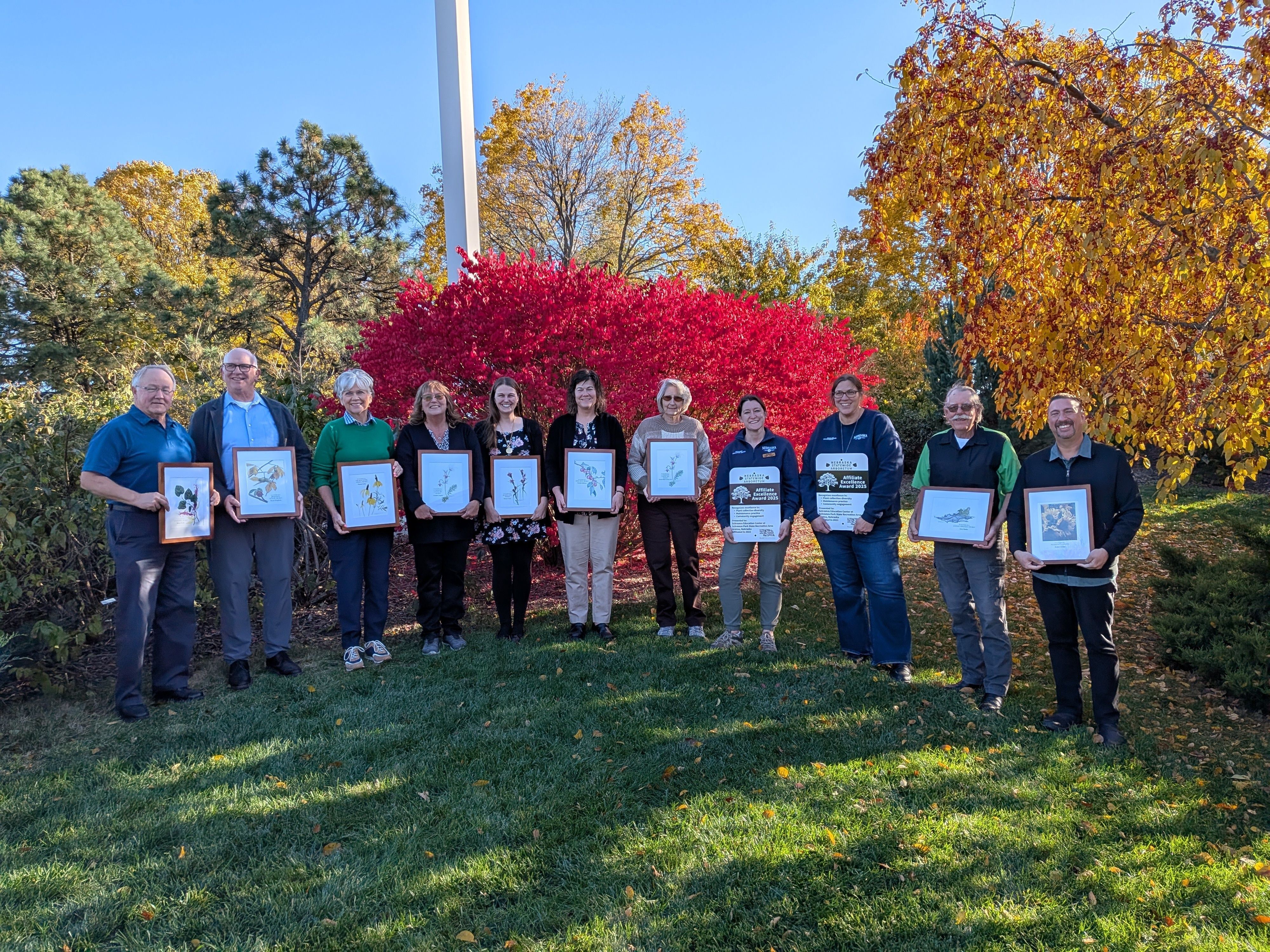 A group of people holding framed awards stand in a semicircle in front of autumnal trees in shades of red and yellow. 