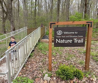 A child walks toward us on a metal bridge beside a sign that says "Welcome to the Snipes Farm and Education Center Nature Trail