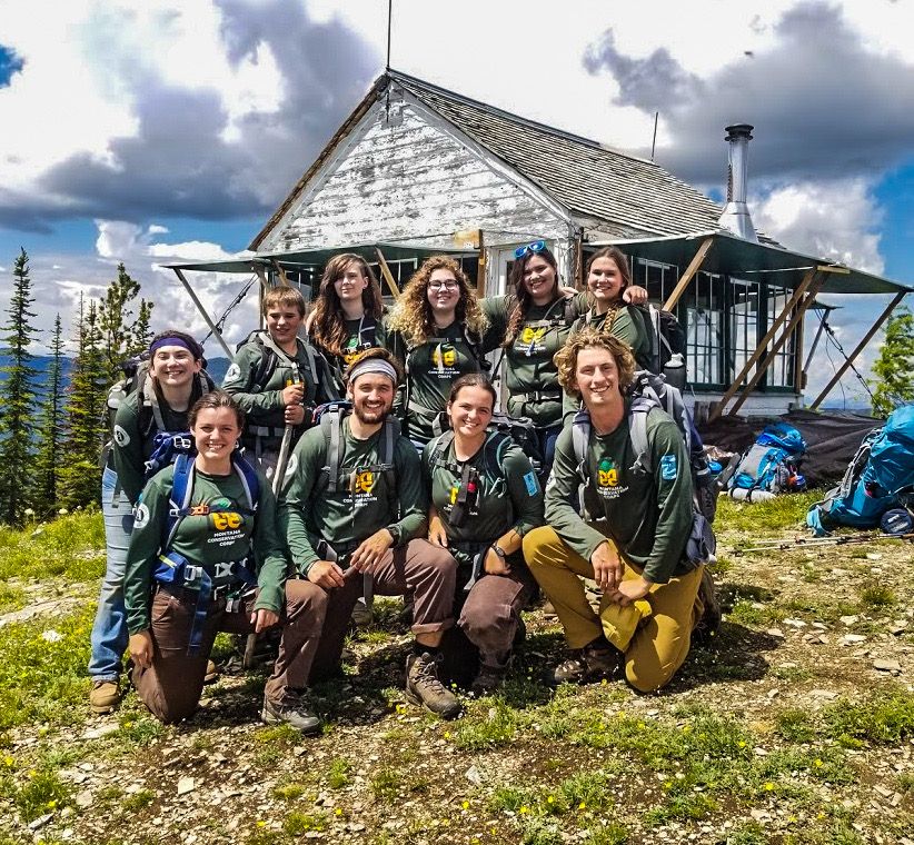 [Image Description: Ten members of an MCC Crew are posed on a mountain top in front of a remote cabin in the wilderness.]