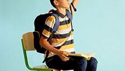 Kid sitting in a classroom chair raising his hand.