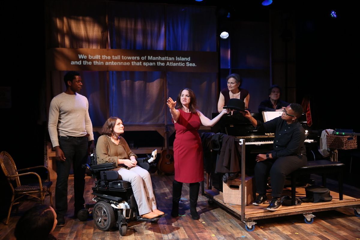 A group shot of the Brecht on Brecht company on stage. The focus is on the woman who is wearing a red dress and she is trying to reach for something while she’s holding a black hat. Everyone else is looking at her.