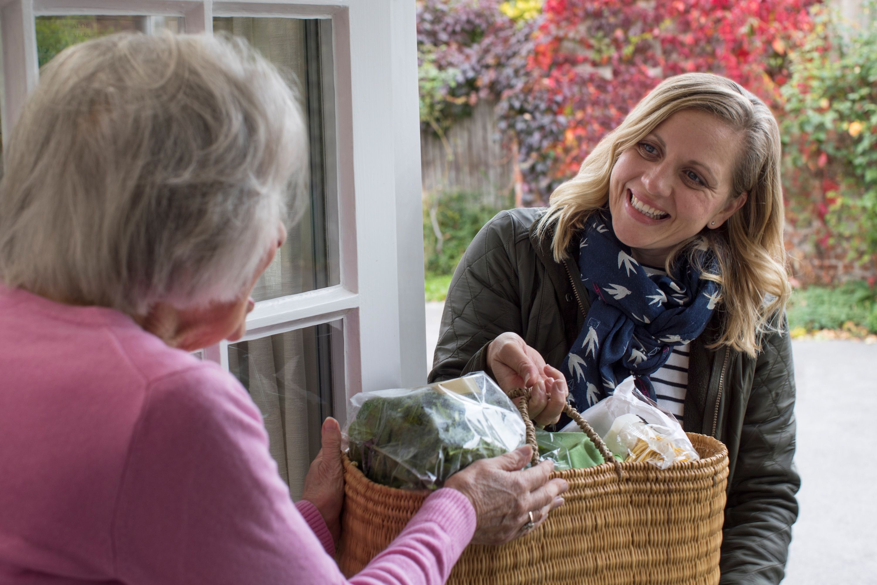 Middle aged woman bringing groceries to older female neighbor, smiling and standing at the front door